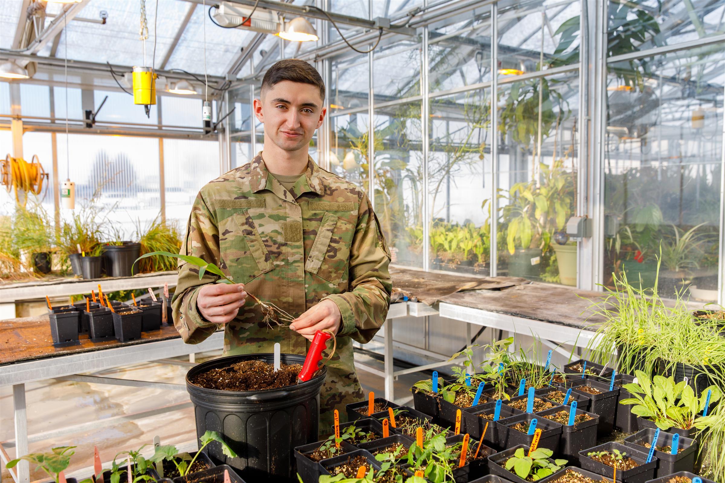 Military student in uniform holding a plant surrounded by plants in pots inside a greenhouse