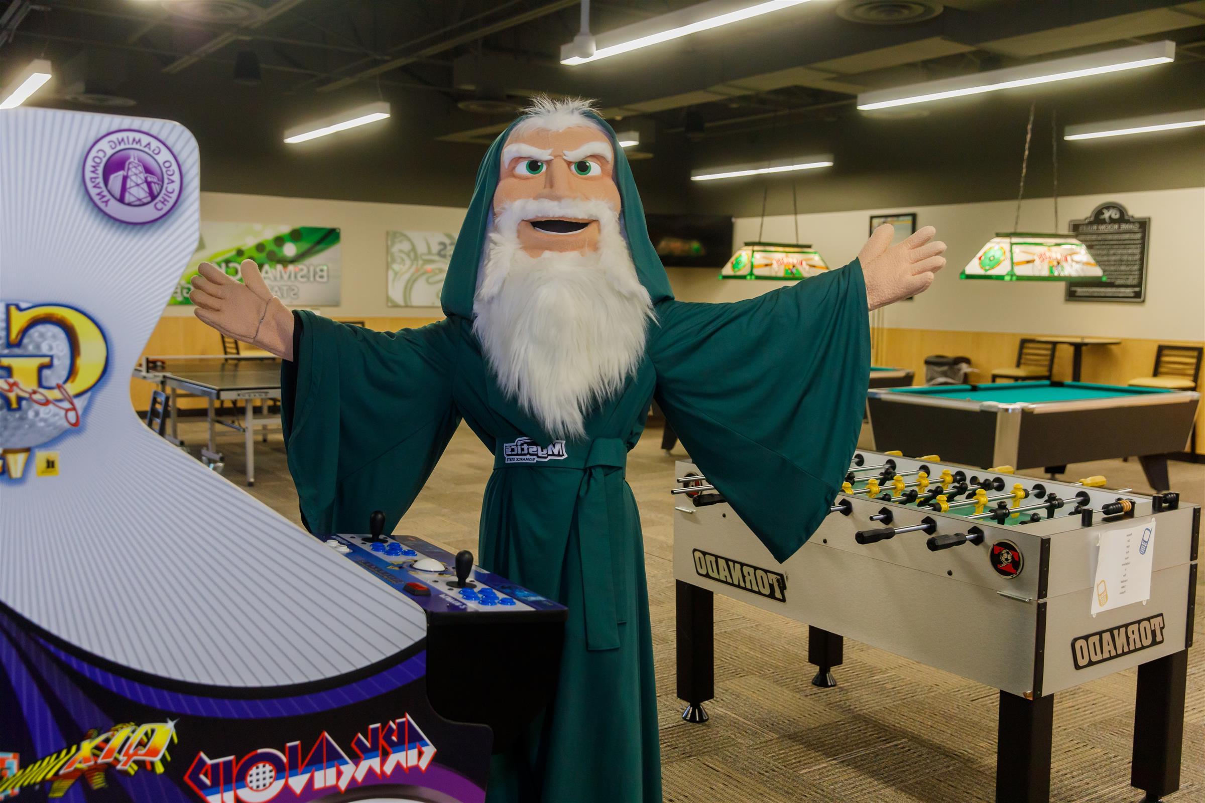 Merlin the Mystic Mascot is holding his arms open wide in the middle of the BSC game room, showing pool tables, foos ball table, and arcade games